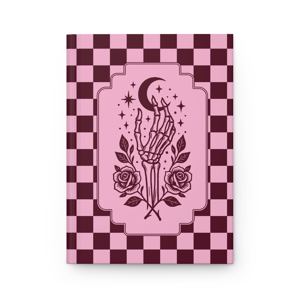 A hardcover notebook or journal with a pink and dark burgundy checkerboard cover, featuring a central illustration of a skeleton hand holding a crescent moon and stars, with roses at the base. The book is shown against a plain white background.
