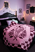 A pink blanket featuring a gothic-style design with a skeleton hand holding roses under a crescent moon and stars, framed by a dark pink and burgundy checkerboard border.