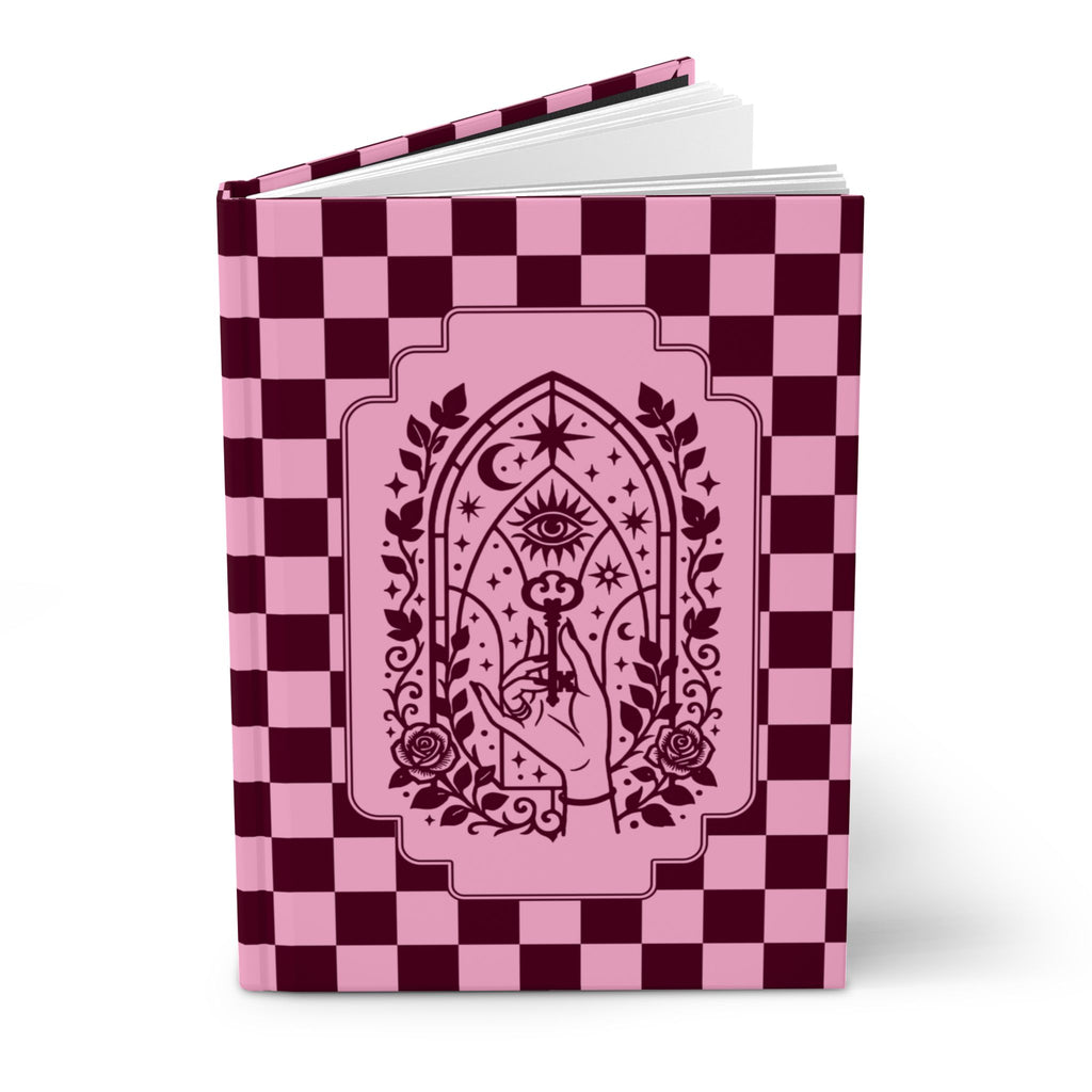 A hardcover notebook or journal standing upright on a white background. The cover features a pink and dark burgundy checkerboard pattern with a central pink arched illustration containing an eye, stars, crescent moon, roses, and a hand holding a wand.