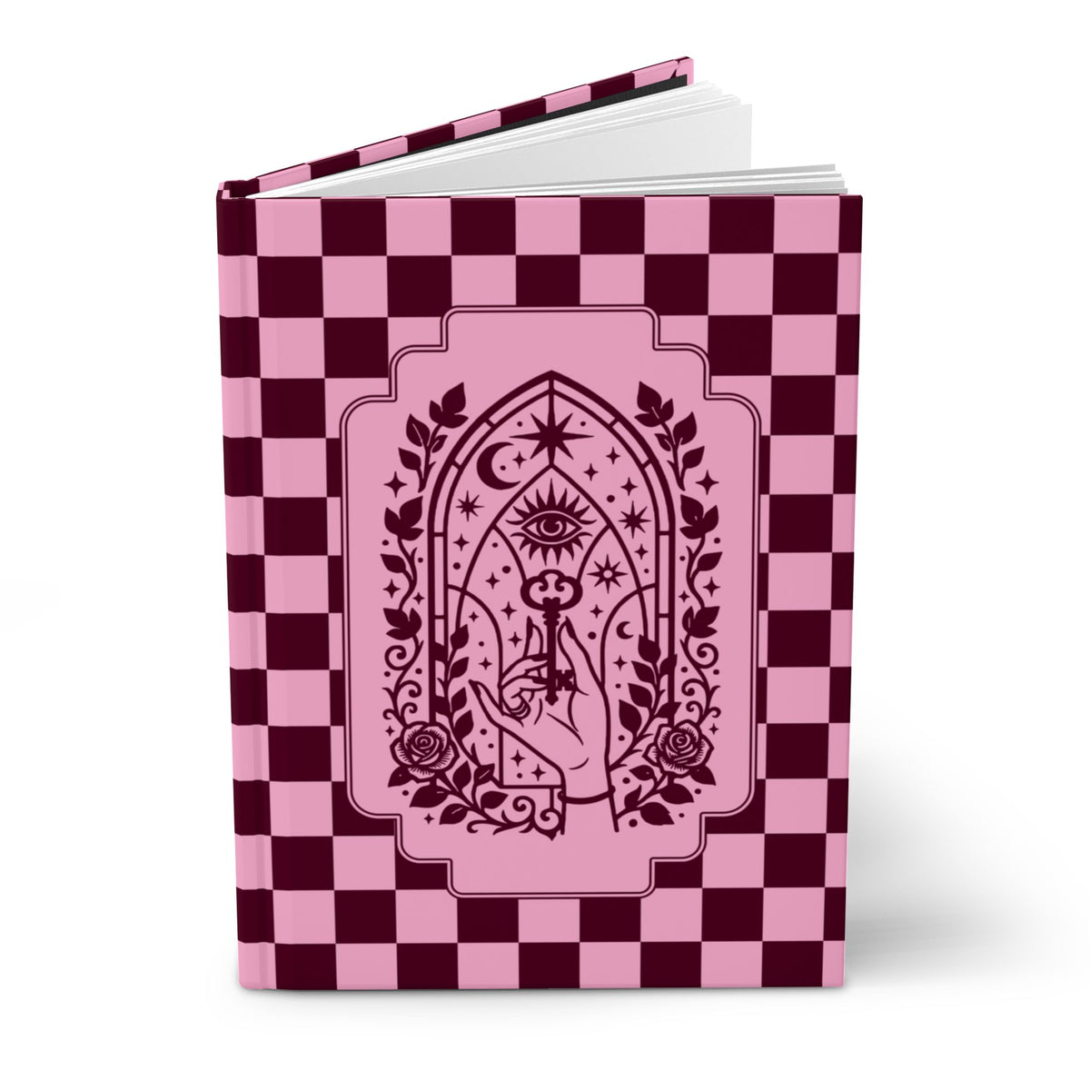 A hardcover notebook or journal standing upright on a white background. The cover features a pink and dark burgundy checkerboard pattern with a central pink arched illustration containing an eye, stars, crescent moon, roses, and a hand holding a wand.