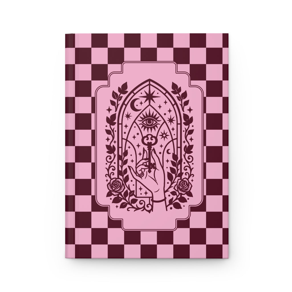 A hardcover notebook or journal standing upright on a white background. The cover features a pink and dark burgundy checkerboard pattern with a central gothic-style illustration of a hand holding a rose under an all-seeing eye and crescent moon motif.