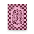 A hardcover notebook or journal standing upright on a white background. The cover features a pink and dark burgundy checkerboard pattern with a central gothic-style illustration of a hand holding a rose under an all-seeing eye and crescent moon motif.
