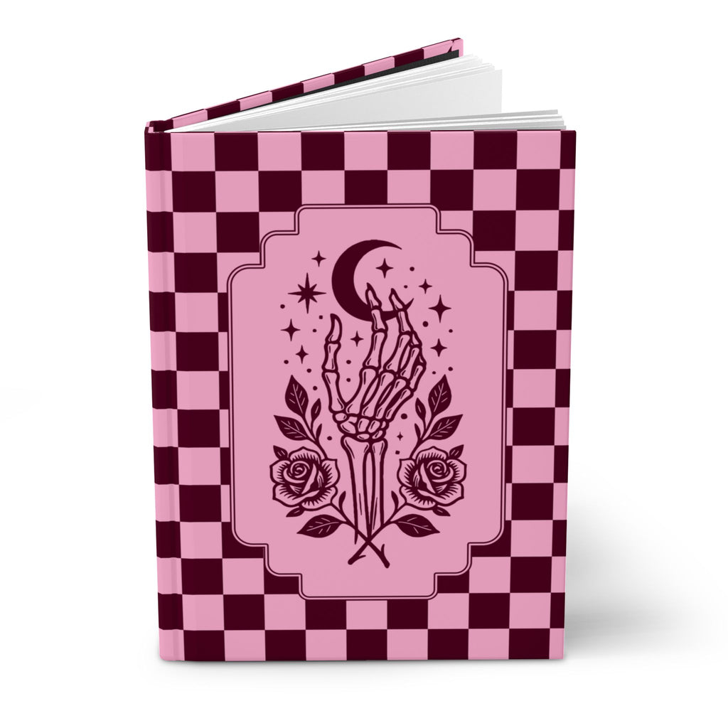 A hardcover notebook or journal with a pink and dark burgundy checkerboard cover, featuring a central illustration of a skeletal hand holding a crescent moon and stars, with roses at the bottom, shown on a white background.