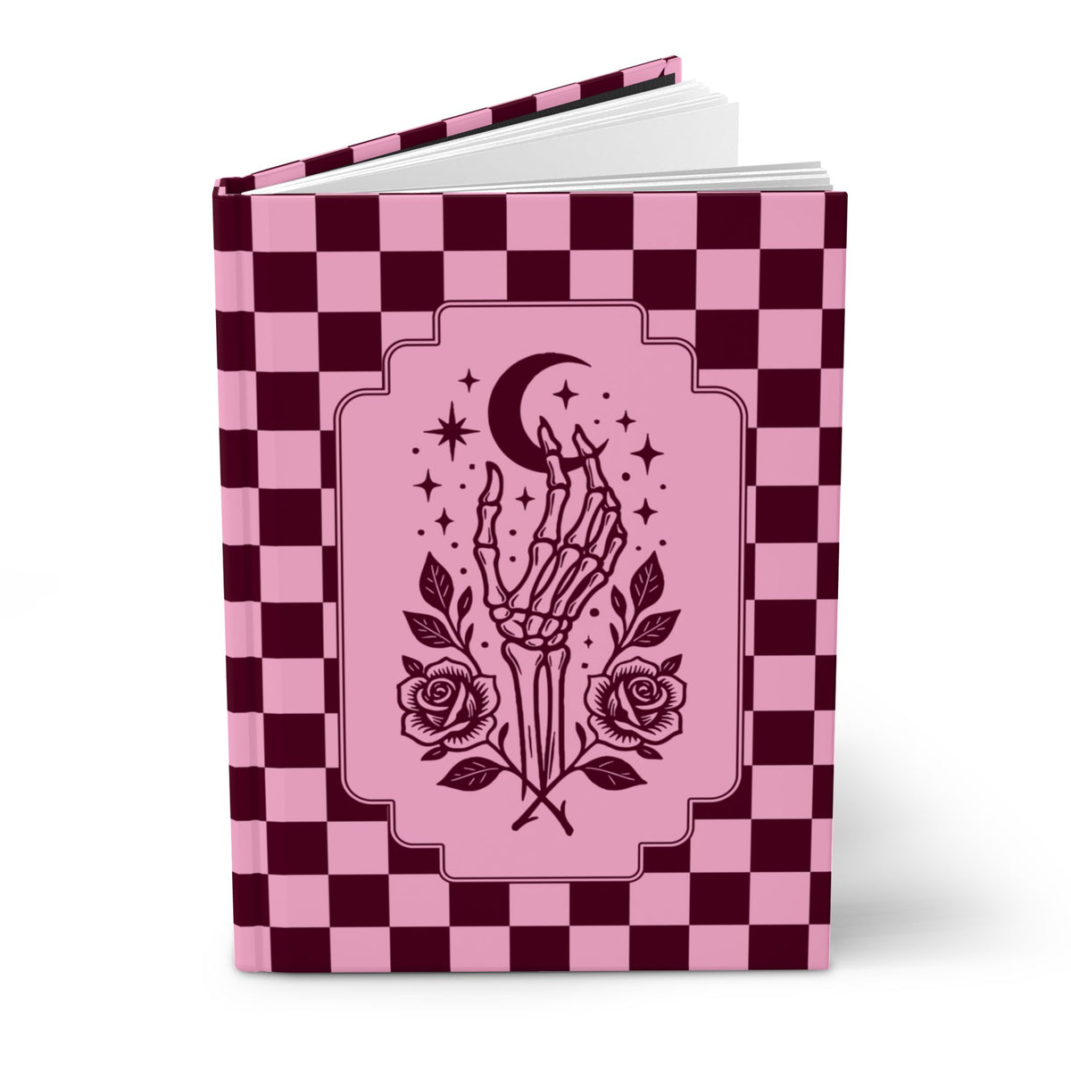 A hardcover notebook or journal with a pink and dark burgundy checkerboard cover, featuring a central illustration of a skeletal hand holding a crescent moon and stars, with roses at the bottom, shown on a white background.