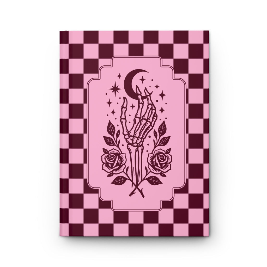 A hardcover notebook or journal with a pink and dark burgundy checkerboard cover, featuring a central illustration of a skeletal hand holding a crescent moon and stars, with roses at the bottom, shown on a white background.