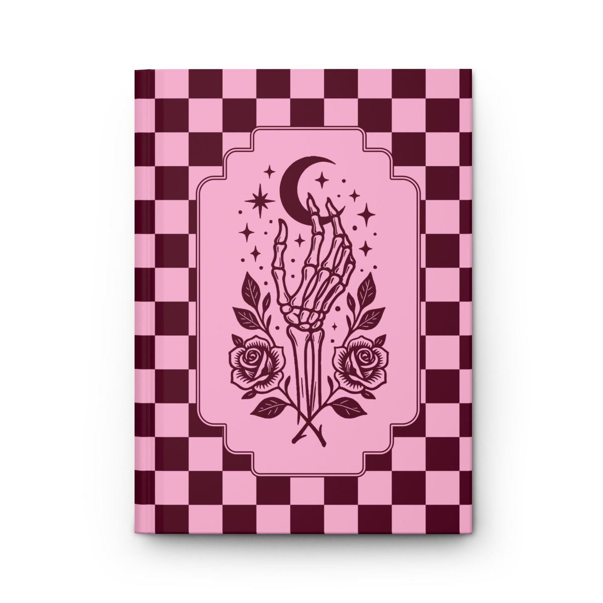 A hardcover notebook or journal with a pink and dark burgundy checkerboard cover, featuring a central illustration of a skeletal hand holding a crescent moon and stars, with roses at the bottom, shown on a white background.