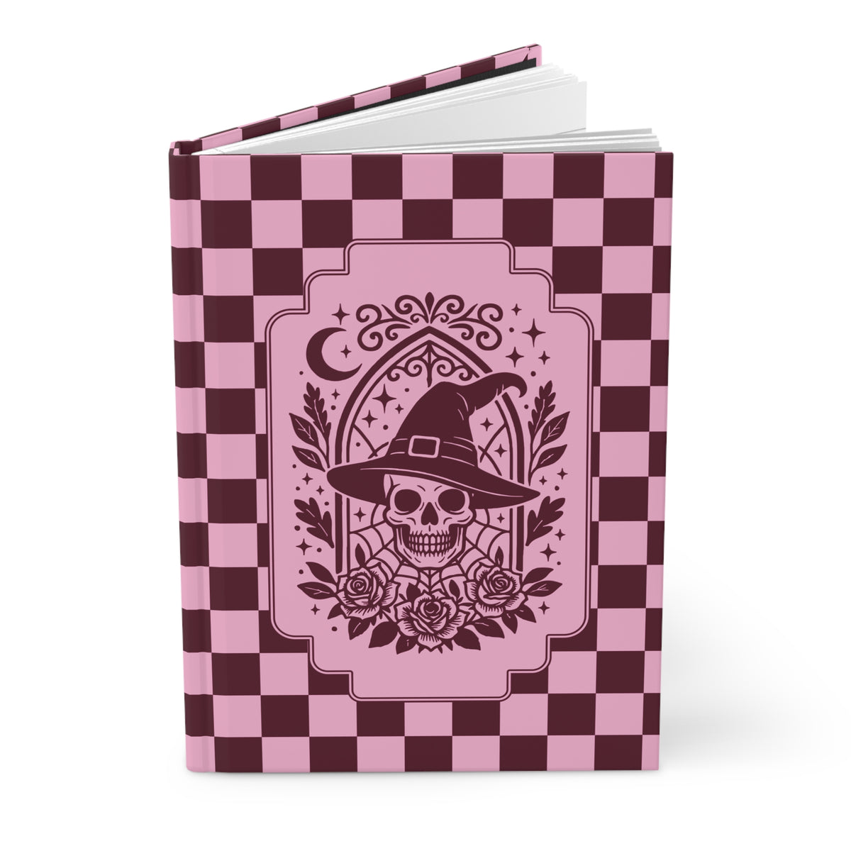 A hardcover notebook with a pink and dark burgundy checkerboard cover, featuring a central graphic of a skull wearing a witch's hat surrounded by roses and a crescent moon.