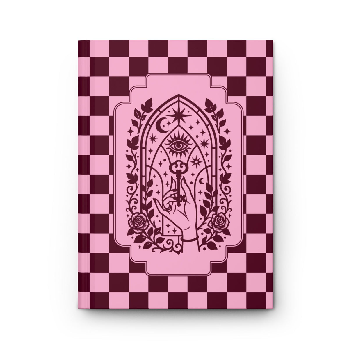 A hardcover notebook or journal standing upright on a white background. The cover features a pink and dark burgundy checkerboard pattern with a central pink arched illustration containing an eye, stars, crescent moon, roses, and a hand holding a wand.