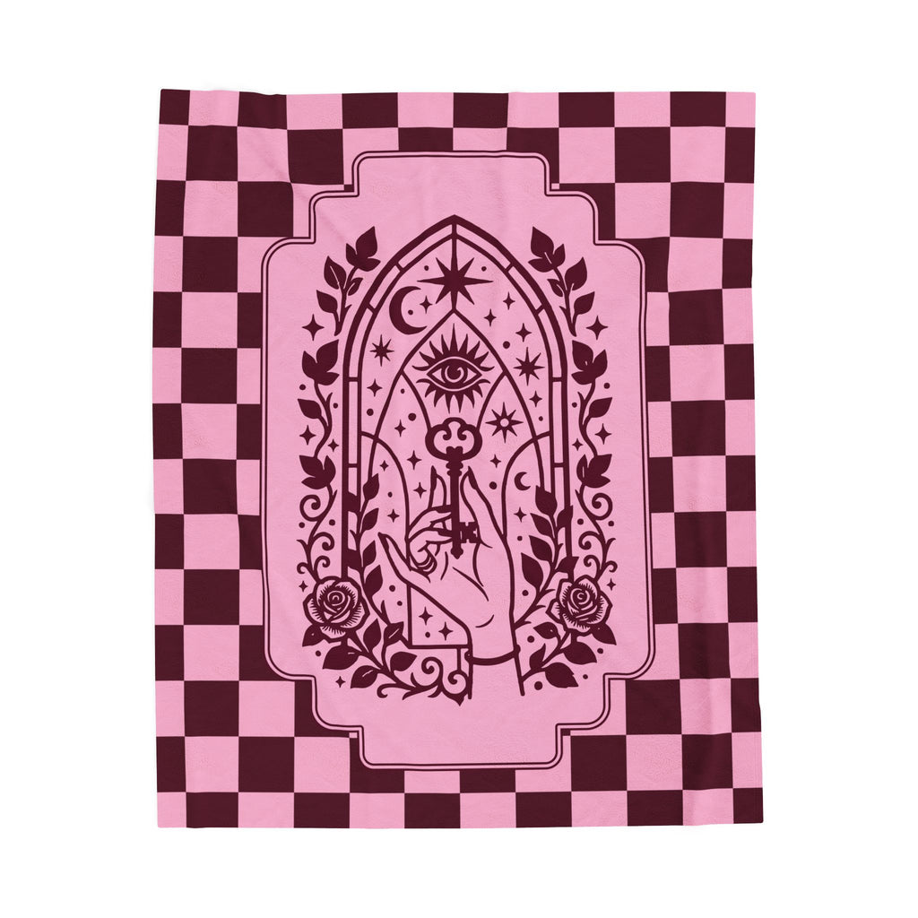 A blanket, featuring a dark burgundy graphic of a hand holding a key in front of an all-seeing eye and rose motif, set against a pink and burgundy checkerboard background.