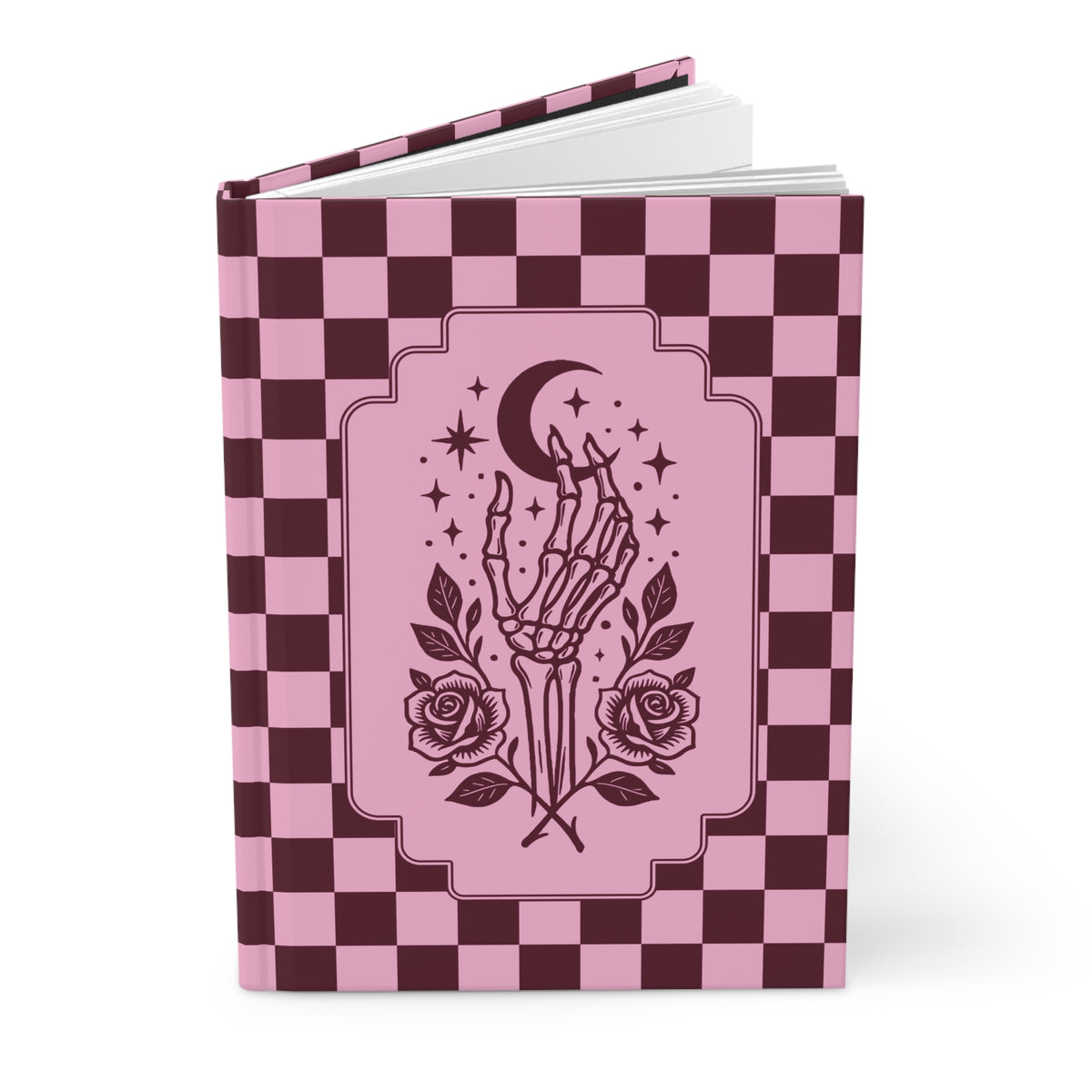 A hardcover notebook or journal with a pink and dark burgundy checkerboard cover, featuring a central illustration of a skeleton hand holding a crescent moon and stars, with roses at the base. The book is shown open against a plain white background.