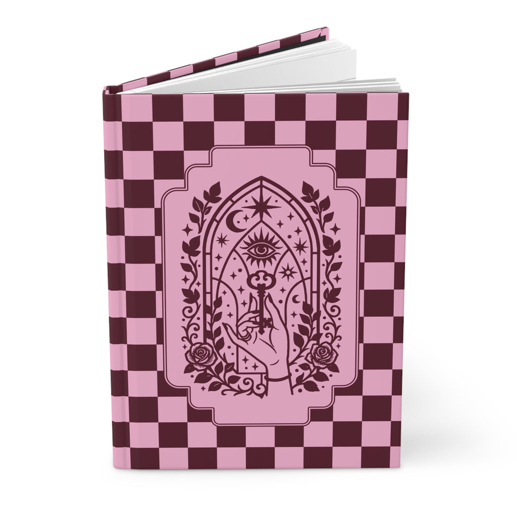 A hardcover notebook or journal standing upright on a white background. The cover features a pink and dark burgundy checkerboard pattern with a central gothic-style illustration of a hand holding a rose under an all-seeing eye and crescent moon motif.