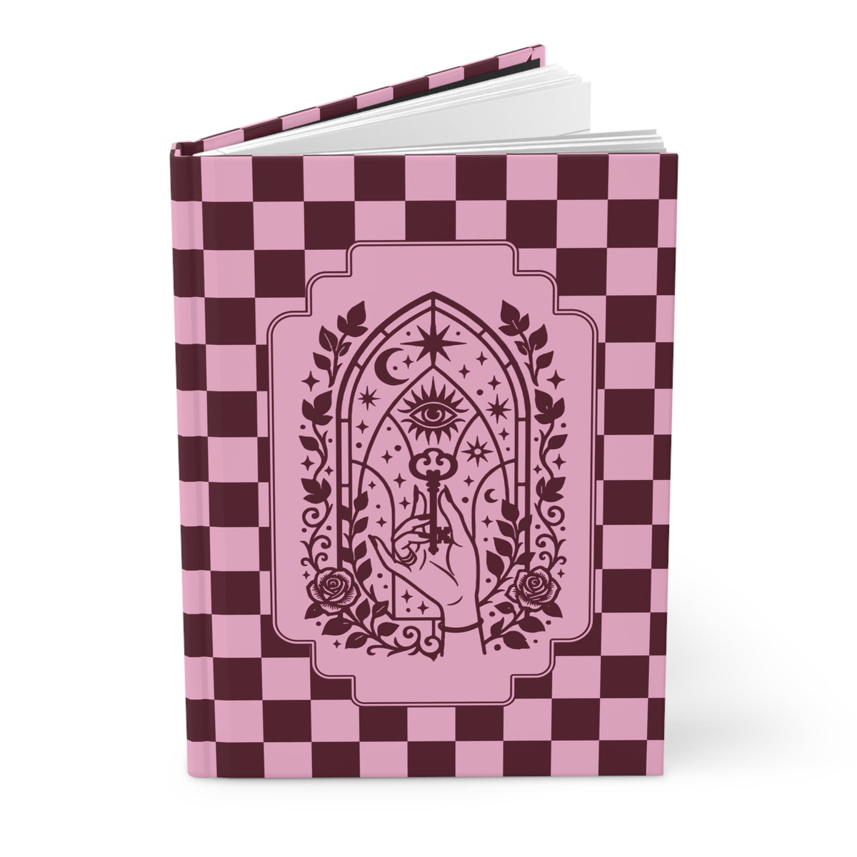 A hardcover notebook or journal standing upright on a white background. The cover features a pink and dark burgundy checkerboard pattern with a central gothic-style illustration of a hand holding a rose under an all-seeing eye and crescent moon motif.