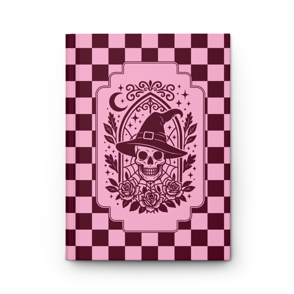 A hardcover notebook or journal with a pink and dark burgundy checkerboard cover, featuring a central graphic of a skull wearing a witch's hat with roses and a crescent moon.

