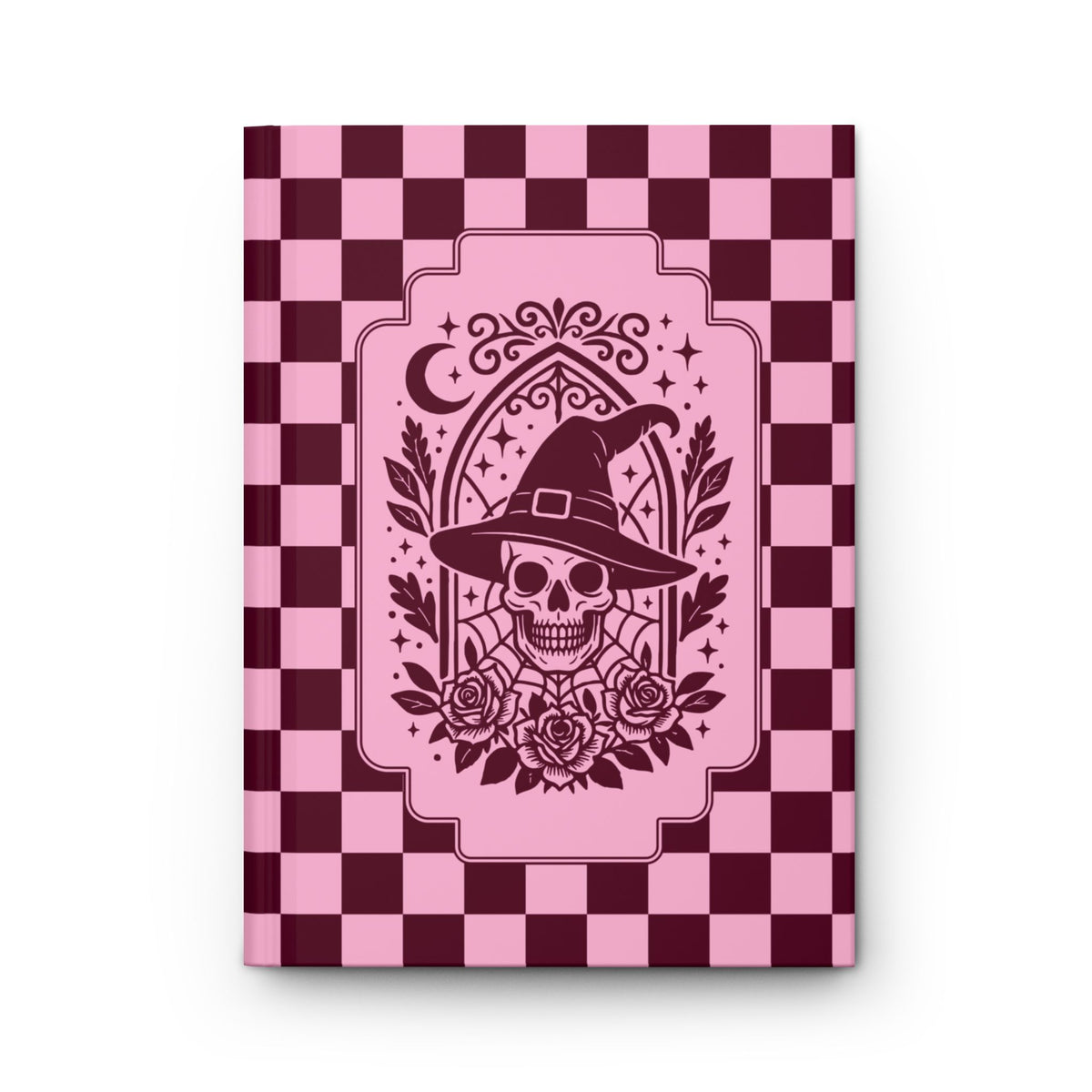 A hardcover notebook or journal with a pink and dark burgundy checkerboard cover, featuring a central graphic of a skull wearing a witch's hat with roses and a crescent moon.

