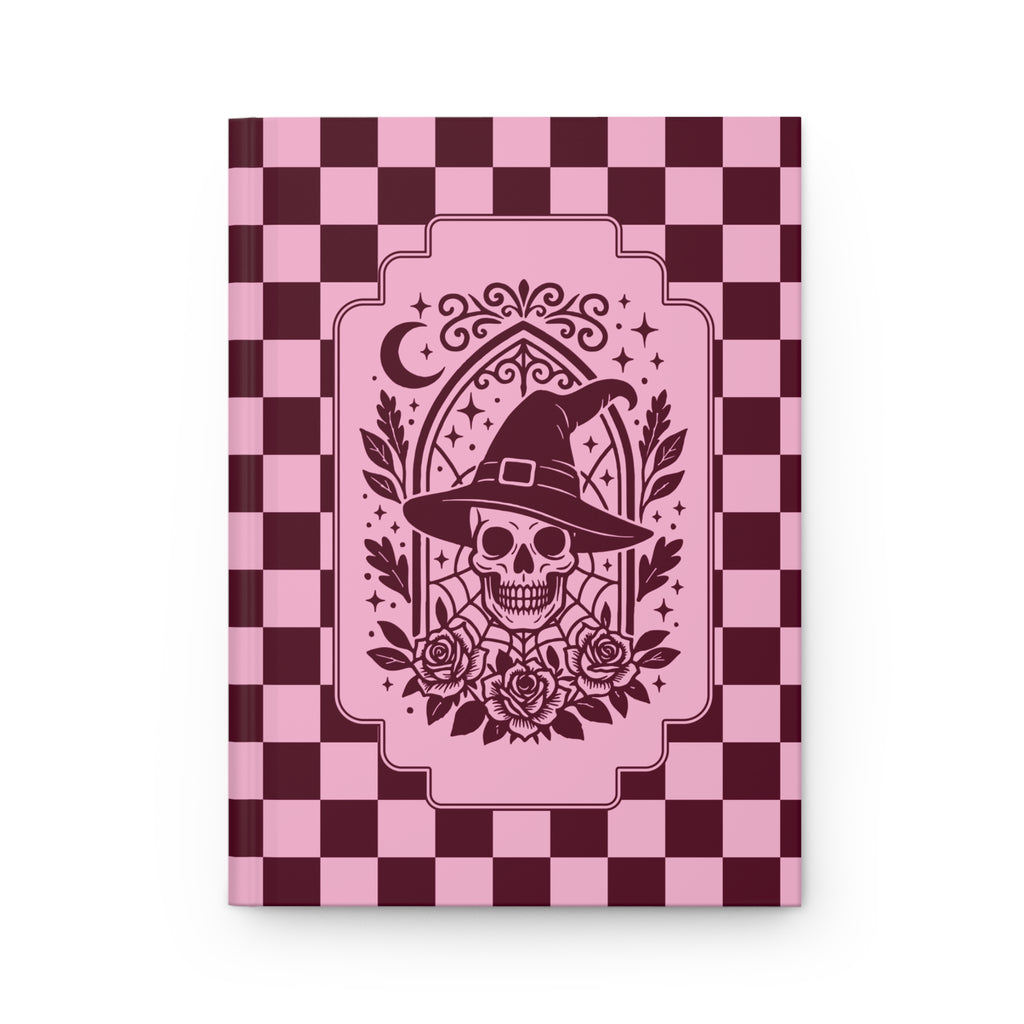 A hardcover notebook with a pink and dark burgundy checkerboard cover, featuring a central graphic of a skull wearing a witch's hat surrounded by roses and a crescent moon.