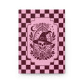 A hardcover notebook with a pink and dark burgundy checkerboard cover, featuring a central graphic of a skull wearing a witch's hat surrounded by roses and a crescent moon.