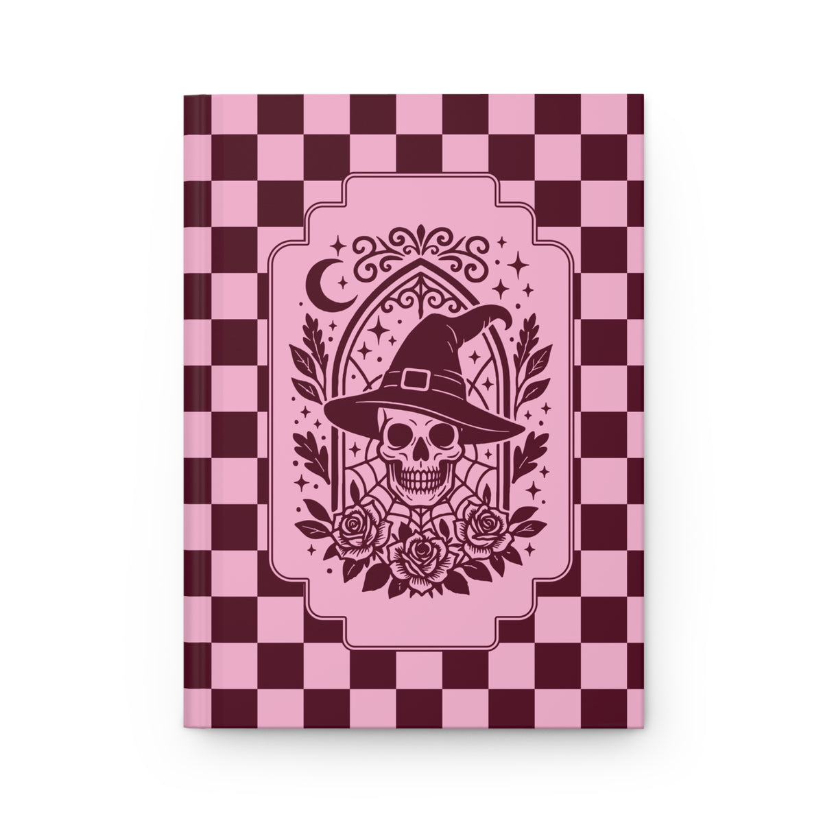 A hardcover notebook with a pink and dark burgundy checkerboard cover, featuring a central graphic of a skull wearing a witch's hat surrounded by roses and a crescent moon.