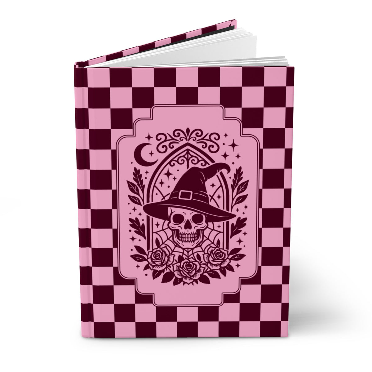 A hardcover notebook or journal with a pink and dark burgundy checkerboard cover, featuring a central graphic of a skull wearing a witch's hat with roses and a crescent moon.