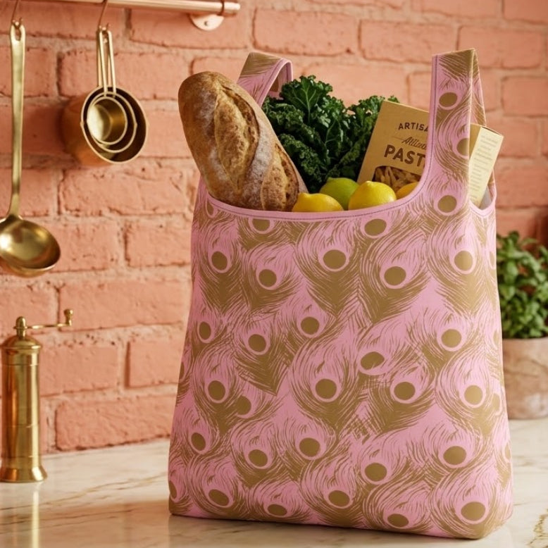 A pink reusable shopping bag with a gold peacock-feather pattern sits on a marble kitchen counter, filled with groceries like a loaf of bread, greens, lemons, and pasta against a terracotta brick wall with hanging copper kitchen tools.