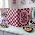 A pink and dark red checkerboard tablet case with a witch skull graphic, displayed on a light-colored wooden dresser surrounded by gothic-themed decor.